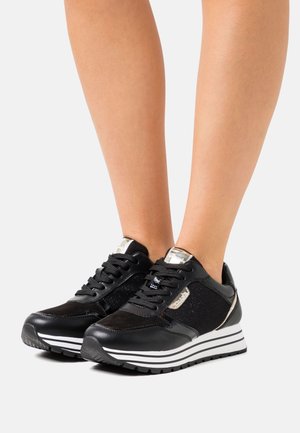 Nohy v černých platformových teniskách s bílými pruhovanými podrážkami a kovově stříbrnými doplňky na světlém pozadí.