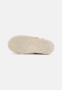 Skoinnersåle i beige gummi med ett zigzag-mönster, med logotypen "UGG." Designen är platt och funktionell för bra grepp.
