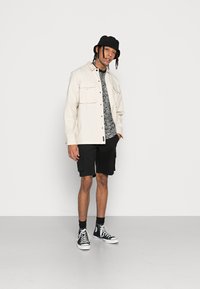Helles beiges Hemd mit Knöpfen, darunter ein schwarzes T-Shirt mit Muster, schwarze Cargo-Shorts, ein schwarzer Bucket Hat und schwarze High-Top-Sneaker.