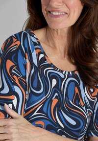 Multikleurige top met een draaiend patroon in blauw, oranje en wit tegen een donkere achtergrond; zachte stof met een ronde halslijn.