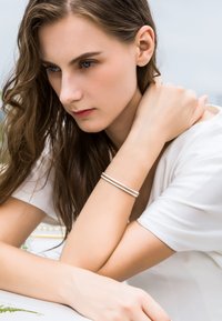 Pulsera delicada de plata con hilos blancos entrelazados, con un acento dorado texturizado. Se lleva en un brazo descubierto, sobre un fondo claro.