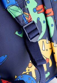 Detailný záber nastaviteľného popruhu na batohu tmavomodrej farby s dinosaurovým vzorom v modrej, zelenej, žltej a červenej na textilnom pozadí.