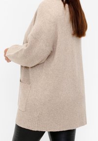 Beżowy sweter z dzianiny z długimi rękawami, prążkowanym wykończeniem i boczną kieszenią. Miękka tekstura, luźny krój i długość do połowy uda.