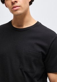 Νέος άνδρας φοράει απλό μαύρο T-shirt με στρογγυλή λαιμόκοψη και τσέπη στο αριστερό στήθος σε ανοιχτό γκρι φόντο.
