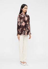 Καφέ πουκάμισο με λουλούδια και κουμπιά, με μακριά μανίκια, συνδυασμένο με κρεμ παντελόνια και λευκές γόβες. Το πουκάμισο έχει μεγάλα μοτίβα από τ roses και λεπτομέρειες με κουμπιά.