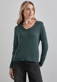 Ciemnozielony sweter dzianinowy z dekoltem w kształcie litery V, długimi rękawami i ściągaczem u dołu. Materiał wydaje się gładki o miękkiej fakturze. Czarne dżinsy dopełniają stylizację.