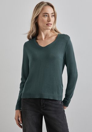 Dunkelgrüner Strickpullover mit V-Ausschnitt, langen Ärmeln und geripptem Saum. Der Stoff wirkt glatt und hat eine weiche Textur. Schwarze Jeans runden das Outfit ab.