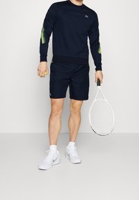 Navy sweatshirt with green mesh accents, navy shorts, and white sneakers. Holding a tennis racket and ball, standing against a plain background.