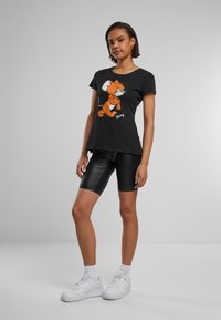 Svart t-skjorte med et tegneseriefigurdesign i oransje, kombinert med svarte skinnende sykkelshorts og hvite joggesko.