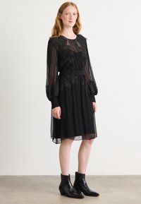 Robe noire avec des manches transparentes en couches, broderie florale sur le corsage, plis, taille cintrée et jupe fluide, associée à des bottines noires.