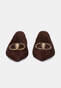 Zapatos planos de ante marrón con punta afilada y herrajes decorativos dorados que presentan una piedra en el centro. Textura suave con un detallado intrincado.