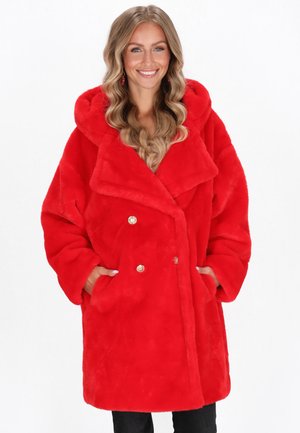 Donna sorridente con lunghi capelli biondo ondulati, indossa un cappotto in faux fur rosso brillante a doppio petto con bottoni dorati e le mani nelle tasche.