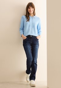Sudadera azul clara con cordones, combinada con jeans de mezclilla oscura y zapatillas blancas. Textura suave y diseño casual. Pose de pie.