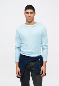 Suéter azul claro de manga longa com decote redondo e punhos canelados, combinado com jeans escuros. Um suéter azul marinho atado à cintura adiciona contraste.