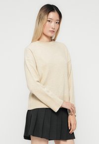 Jeune femme aux cheveux longs portant un pull beige et une jupe plissée noire, se tenant devant un fond blanc uni.