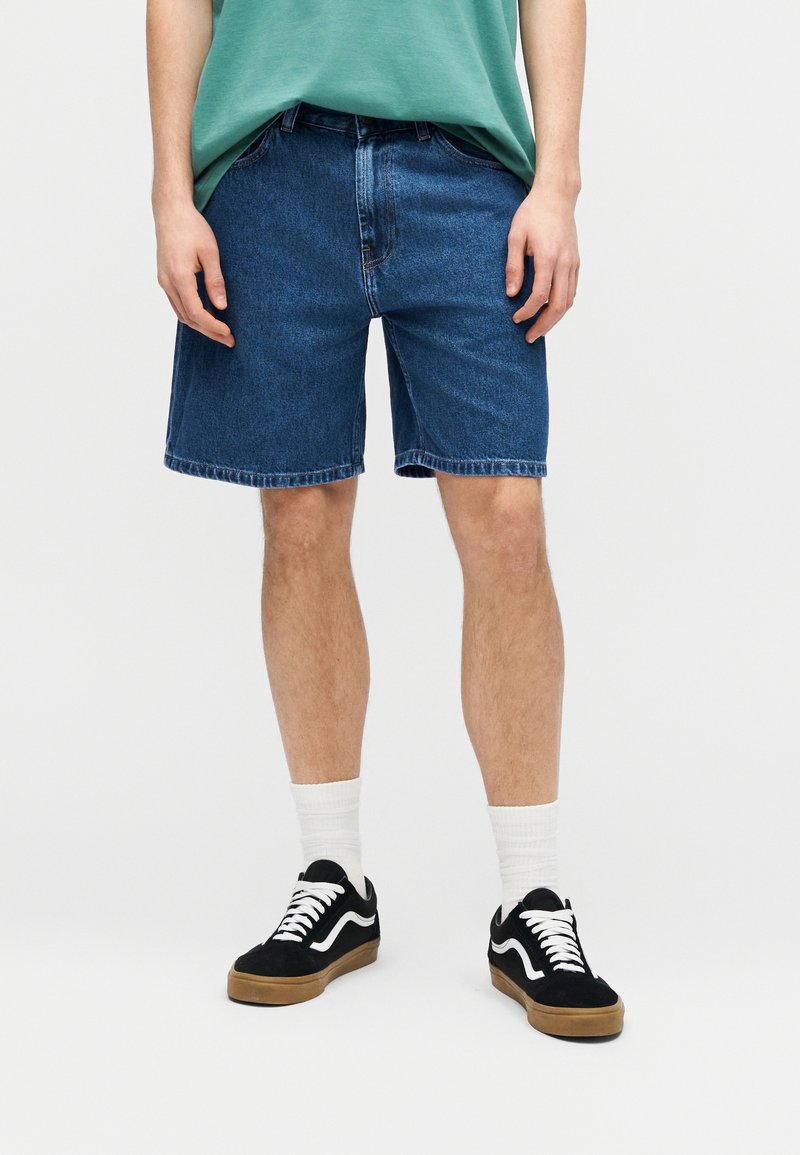 Persona que lleva pantalones cortos de mezclilla azul, zapatillas negras y blancas, calcetines blancos y una camiseta verde, de pie frente a un fondo liso.