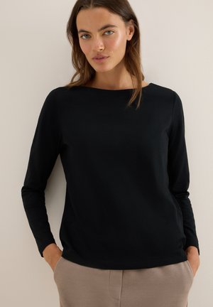 Haut noir à manches longues avec un col rond. Texture lisse et légère flexibilité. Porté de manière décontractée avec les mains dans les poches, sur un fond neutre.