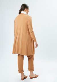 Lange, open voorkant cardigan in zachte camel brei met zijzakken, gedragen over bijpassende tapered pants. Platte schoenen met decoratieve accenten.