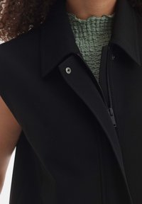 Czarna bezrękawnikowa kurtka z uniesionym kołnierzem i zamkiem błyskawicznym z przodu. Pod spodem teksturowana zielona bluzka z falbaniastym dekoltem.