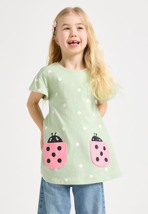 Ung blond pige smilende, iført en mintgrøn polkaprikket kjole med rosa mariehøne-lommer og blå jeans, hænderne bag ryggen.