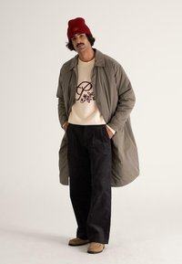 Letthørt, olivengrøn frakke over en cremefarvet sweater med et maroon broderet design, parret med sorte fløjlbukser og brune sko. Rød hue.