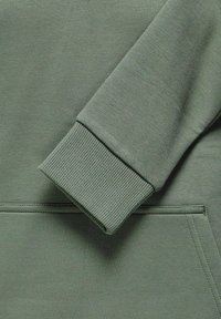 Section de tissu en coton vert pour sweatshirt, avec un poignet côtelé, une couture droite et une ouverture de poche visible en bas.