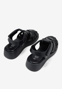 Sandalias negras con un diseño de tiras trenzadas, suela acolchada y plataforma elevada. Cierre ajustable con hebilla. Textura suave y acabado mate.