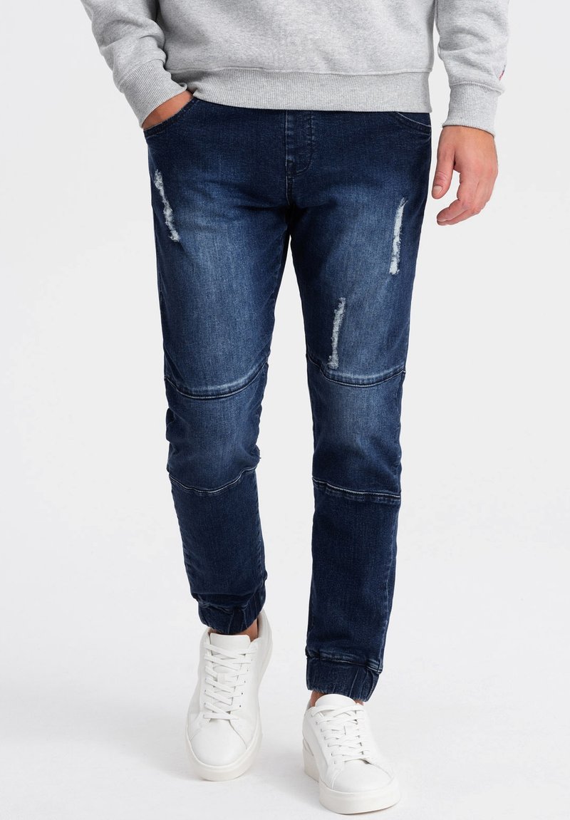 Joggers en denim bleu foncé avec des déchirures et des panneaux au niveau des genoux, dotés d'une finition élastiquée aux chevilles, associés à des baskets blanches.