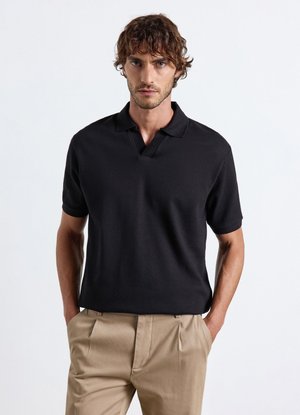 Uomo con capelli castani ricci che indossa una polo nera a maniche corte e pantaloni beige con le mani nelle tasche su sfondo bianco.