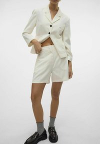 Blazer su misura color crema con bottoni neri, abbinato a pantaloni corti coordinati. Il modello indossa mocassini neri e calzini alla caviglia grigi.