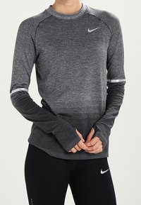 Dlouhý rukáv atletického trička v odstínu šedé s prodyšným materiálem, raglánovými rukávy a reflexními detaily poblíž manžet. Přítomné logo Nike.