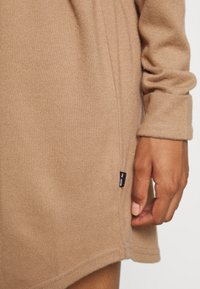 Robe en tissu texturé beige à manches longues. Étiquette de couture latérale notable avec du texte noir. Gros plan sur une main tenant l'ourlet.