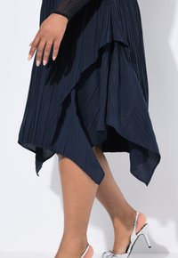 Mujer vestida con una falda asimétrica de tela plisada en color azul marino y zapatos de tacón alto plateados tipo slingback, con la mano descansando al lado de la falda.