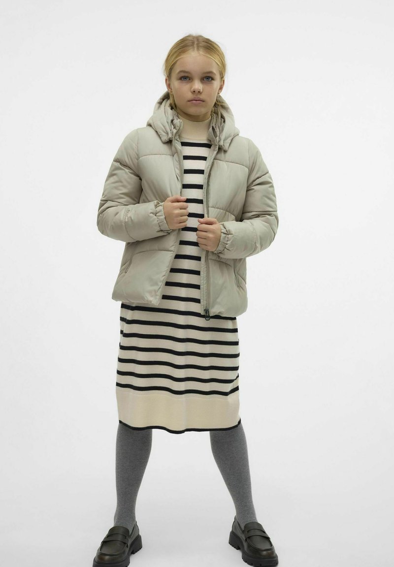 Beige péřová bunda s kapucí s prošívaným designem. Nositá přes šaty s černobílými pruhy a šedé punčocháče s černými loafers.