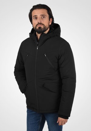 Veste noire isolée avec capuche, dotée de poches poitrine et poches pour les mains zippées, texture lisse et design ajusté. Portée par-dessus un jean bleu.