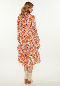 Rochie florală cu un model vibrant portocaliu, roz și alb, cu mâneci lungi și un tiv în mai multe straturi. Modelul poartă cizme bej.
