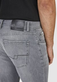 Jean en denim gris avec une patch en cuir noir sur la taille. Comprend deux poches arrière avec des coutures contrastées et une texture lisse.