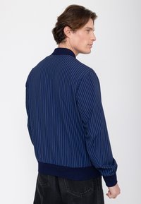 Uomo con capelli castani che indossa una giacca blu navy a righe sottili e pantaloni neri, visto da dietro e leggermente girato di lato.