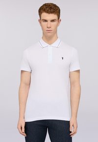 Bílá polokošile s krátkým rukávem, dvoubarevné zapínání na knoflíky, modré akcenty podél límce a malé logo na levé části hrudníku.