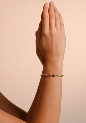 Gold metal bracelet with a twisted rope design, featuring a simple clasp. The hand is positioned with palms together on a soft background.