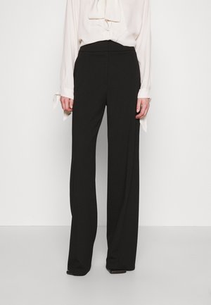 Een vrouw draagt zwarte wijde hoge taillebroeken met een witte blouse met lange mouwen die in de broek is gestopt.