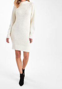 Femme portant une robe-pull en tricot couleur crème à manches longues et des bottines noires, debout devant un fond blanc.