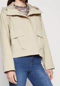 Kvinde iført beige hættetrøje med store forlommer og blå jeans, cropped under skuldrene, stående foran en ensfarvet baggrund.