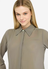 Halenka s dlouhým rukávem v tlumené olivové barvě s knoflíkovým zapínáním, která má velký límec a perlový ozdobný knoflík. Hladká, lehká látka.
