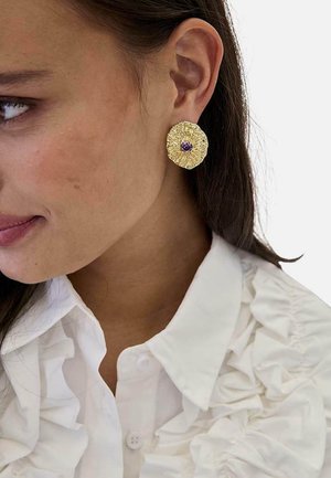 Mujer que lleva un pendiente redondo texturizado de oro con una pequeña piedra púrpura en el centro y un cuello de camisa blanco con volantes.