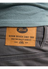 Etiqueta de cuero marrón sobre denim gris, con la inscripción "Petrol Denim Driven Since 1989 Crafting Quality Garments", con detalles de costura visibles.