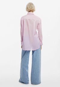 Camicia rosa a maniche lunghe con yoke posteriore e fessure laterali, abbinata a jeans in denim blu a gamba larga. Texture liscia e dettagli del colletto visibili.