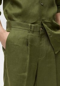 Olive zelený lněný outfit s košilí na knoflíky a plisovanými kalhotami s opaskem. Hladká textura a přizpůsobený střih jsou zřejmé.