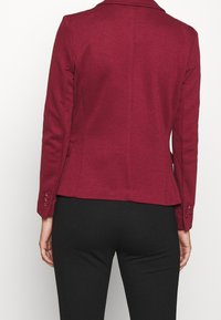 Bordeauxrode aansluitende blazer met een reflecterende kraag, lange mouwen en knoopaccenten op de manchetten. Glad textuur met een getailleerd silhouet.