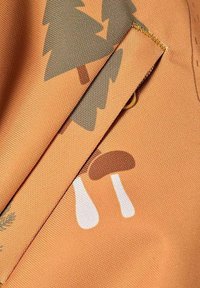 Orange tyg med en strukturerad yta, som har ett tryck av träd och en svampdesign i brunt och vitt. Guldzipperdetalj synlig.